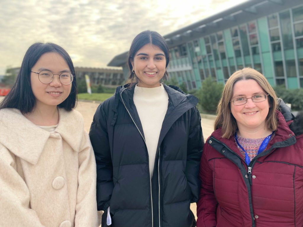 Three women are standing next to each other with a building visible in the background. Yichen wears a white coat and has short black hair, Saamin is the tallest and wears a black coat, Sarah has medium length brown hair and is wearing a red coat