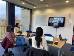 Three people are sat around a table, with a large screen showing other team members on a zoom call